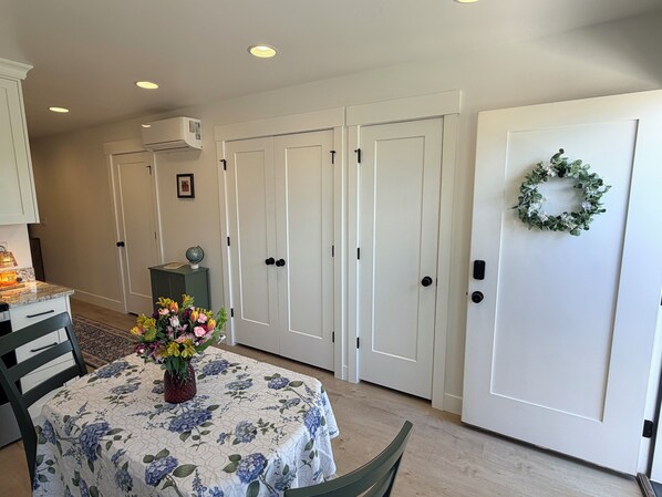 A room with a dining table, chairs, a floral tablecloth, a potted plant, a globe, a small cabinet, and a door with a wreath.