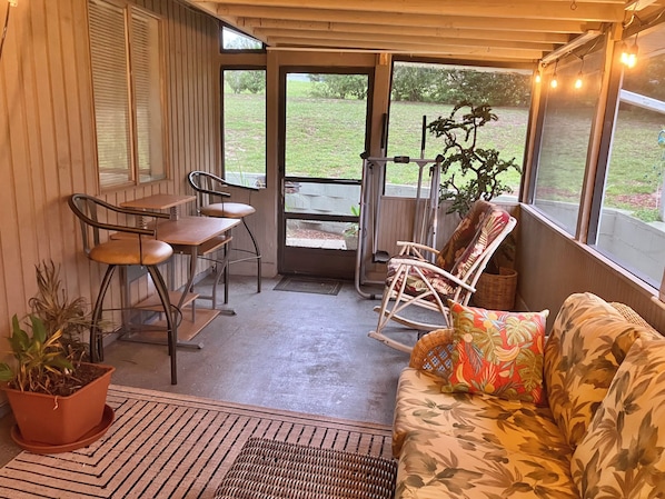 A covered patio area with a wooden table and chairs, a wicker sofa with floral cushions, and a potted plant.