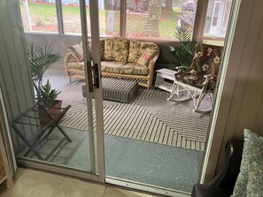 Ein Zimmer mit einer Schiebeglastür, die zu einer Terrasse im Freien führt. Innenbereich mit einem Rattansofa mit floralen Kissen, einem kleinen weißen Tisch und einem Schaukelstuhl.