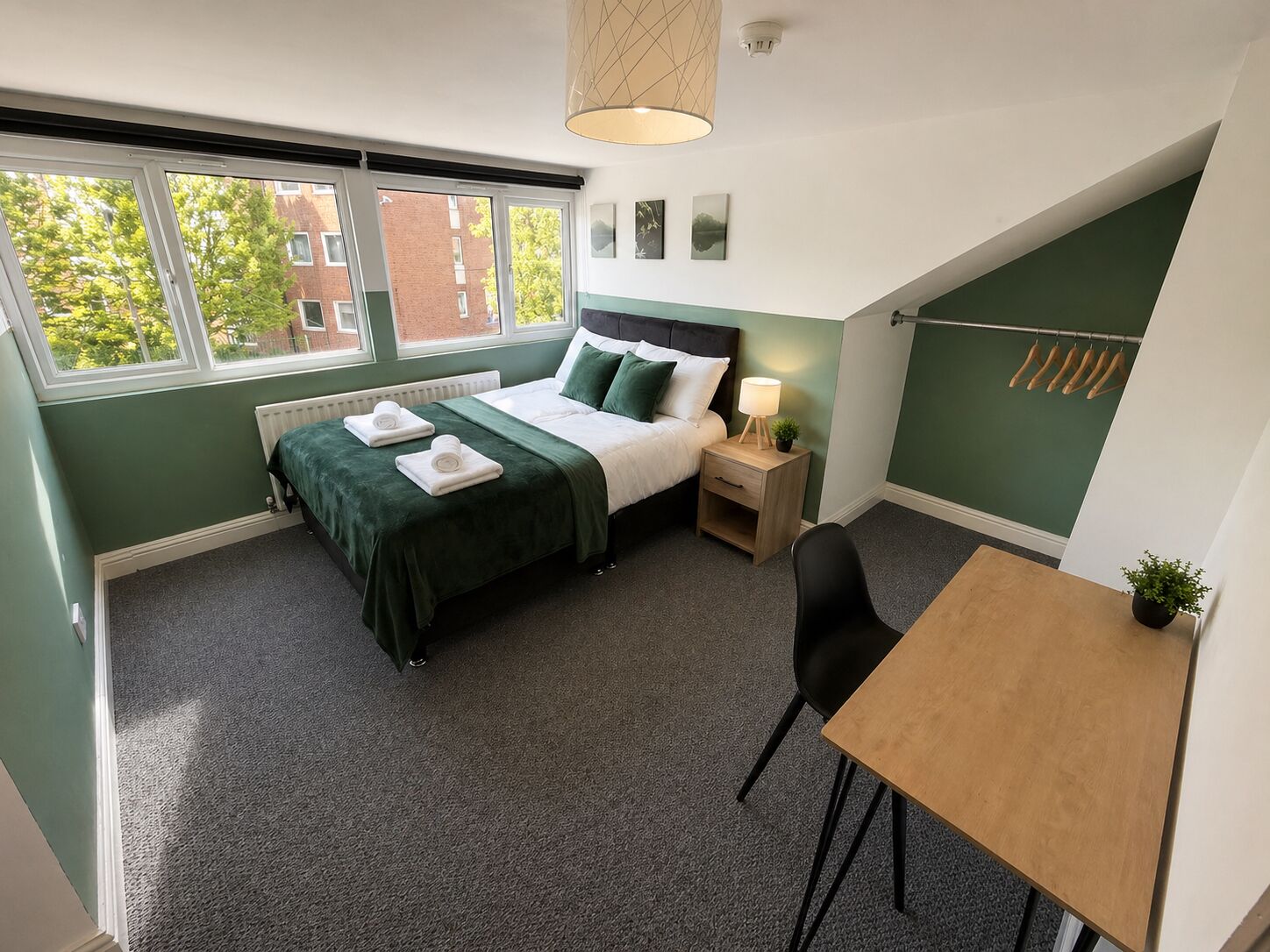 Una camera da letto con un letto, comodino, lampada, sedia e vista sugli edifici attraverso la finestra.