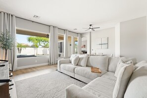 Ein modernes Wohnzimmer mit einer Eckcouch, einem großen Fenster mit Vorhängen, einem Deckenventilator und Blick ins Freie.
