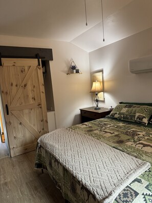 A bedroom with a wooden door, a bed with patterned bedding, a bedside lamp, and a wall-mounted air conditioner.