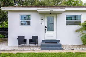 A small, single-story building with a white exterior, numbered 928, featuring two windows, a door with a glass panel, and two black chairs outside. - Cozy Palm Beach Stay 5min Downtown Pets OK (West Palm Beach)
