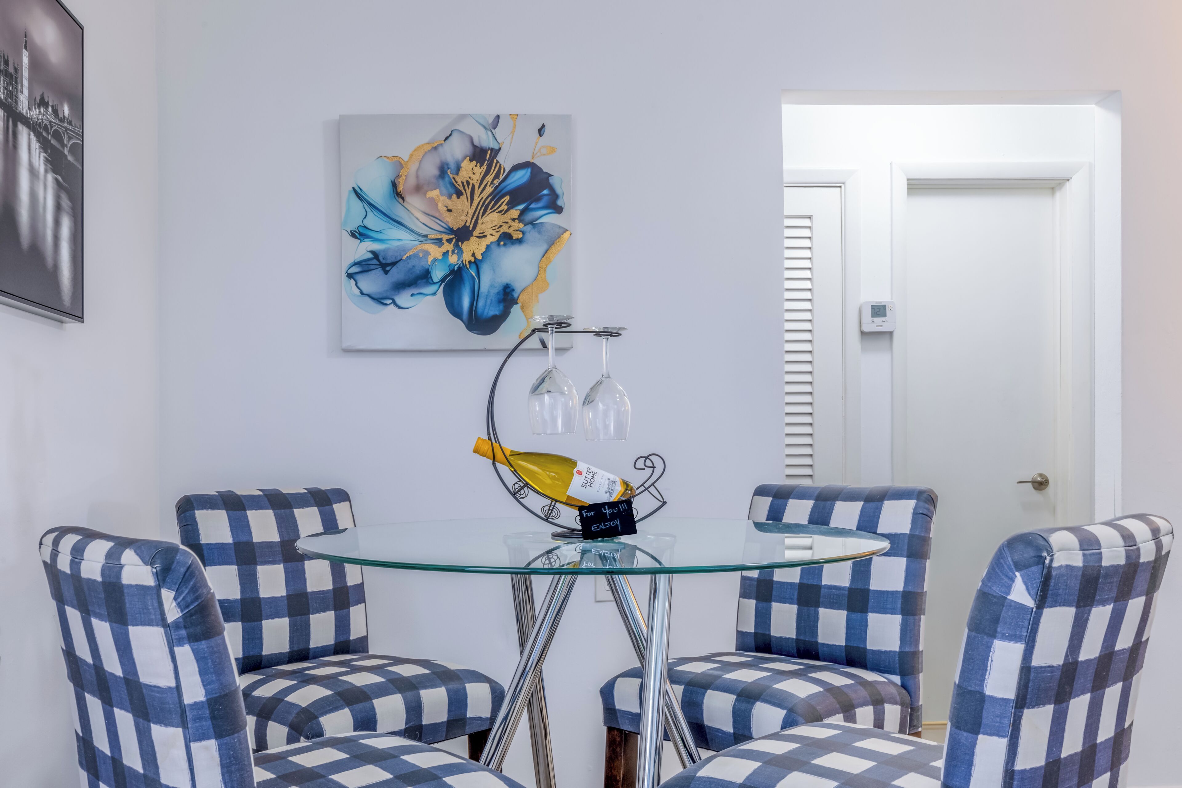 Un comedor con mesa de vidrio, sillas a cuadros, un cuadro floral y una puerta con sensor.