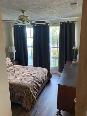 Ein Schlafzimmer mit einem Bett, einem Schreibtisch, einer Lampe, einem Deckenventilator und einem Fenster mit Vorhängen.