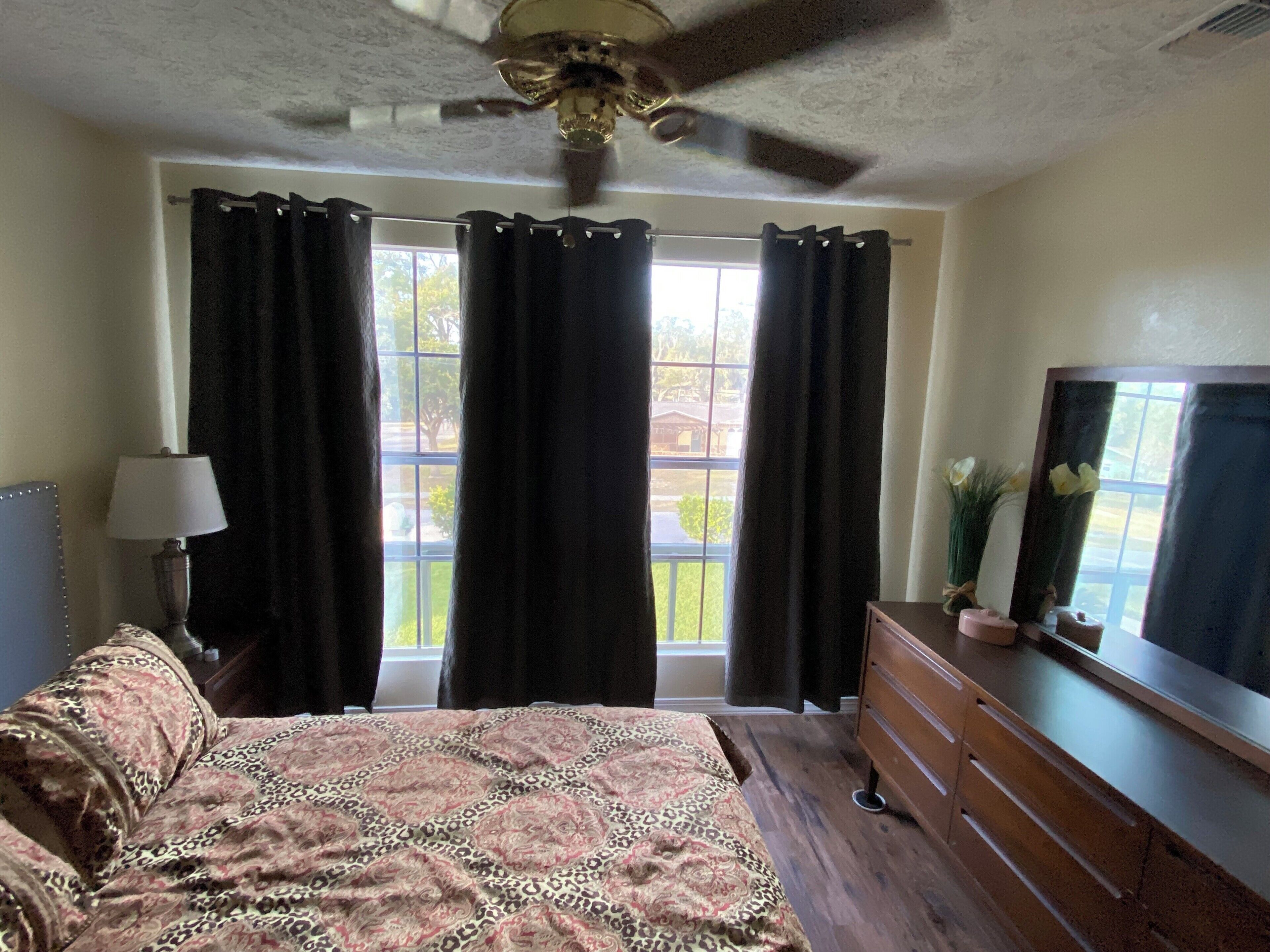 Une chambre à coucher comprenant un lit, une commode, un miroir, un ventilateur de plafond et une fenêtre avec des rideaux.