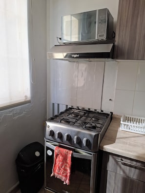 Una cocina con estufa a gas, microondas, un cesto de basura y una ventana.