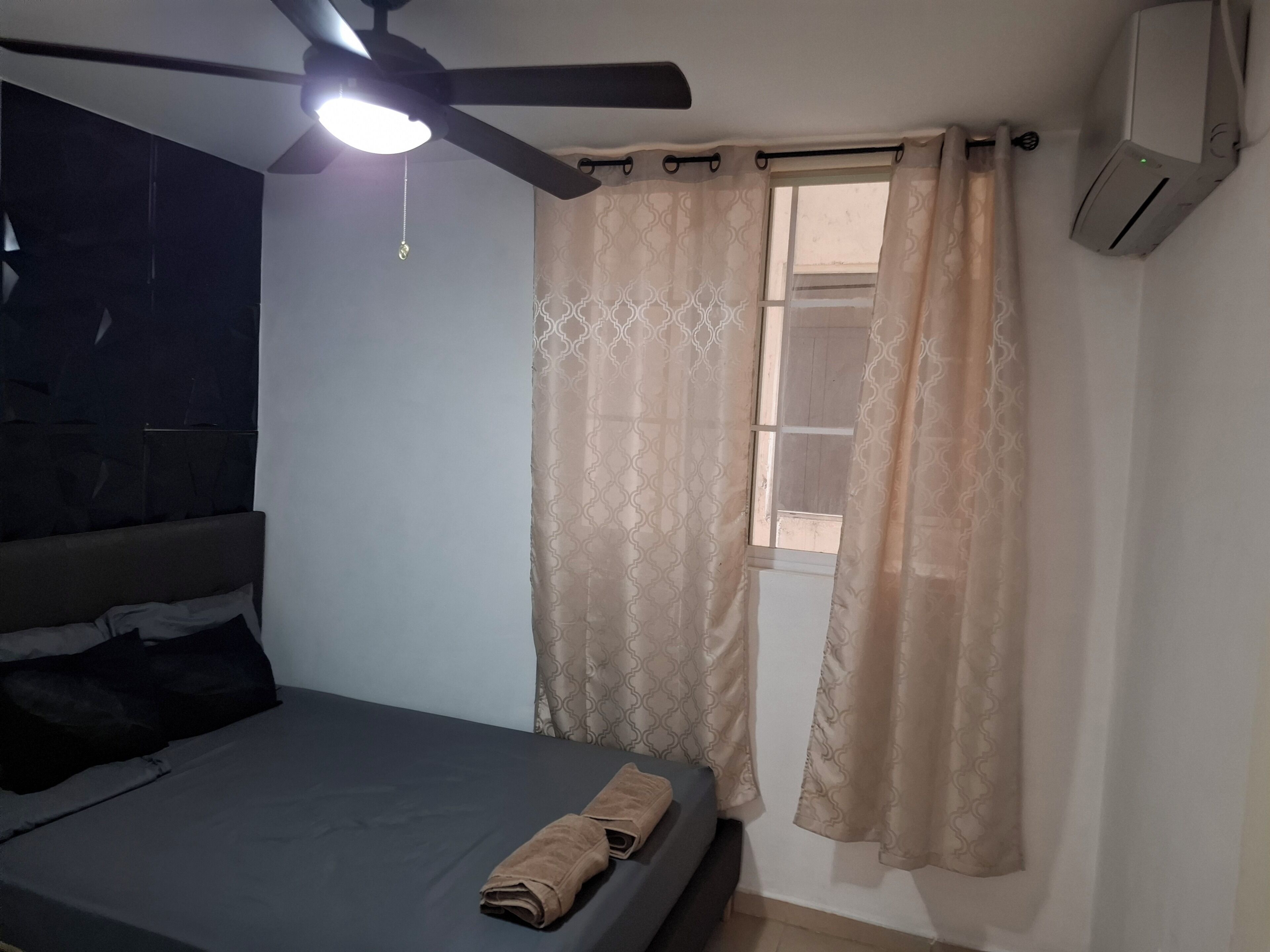 Ein Schlafzimmer mit Bett, Deckenventilator, Fenster mit Vorhängen und Klimaanlage.