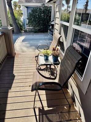 A wooden deck with a chair and two potted plants, a small table, and a window overlooking a garden. - Palm Creek Resorts & Residences Charming 2-Bedroom 2-Bth Village House. (Casa Grande)