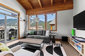 A modern living room with a sofa, coffee table, and a view of a mountainous landscape through large windows.