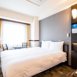 Ein Hotelzimmer mit einem großen Bett, einem kleinen Tisch mit Stühlen, einem Fenster mit Vorhängen und einem an der Wand montierten Fernseher.