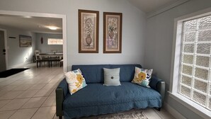 A living room with a blue sofa, two framed pictures, a dining area, and a large window. - Private Gated Montrose Cottage Near FIFA & Downtown (Houston)