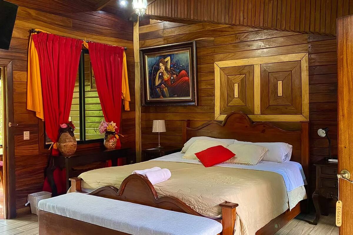 A wooden-paneled room with a bed, a nightstand, a lamp, and a painting on the wall.