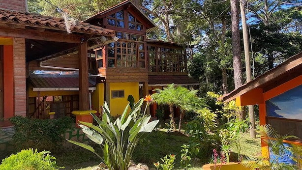 Uma casa de dois andares com varanda, cercada por vegetação exuberante e árvores.