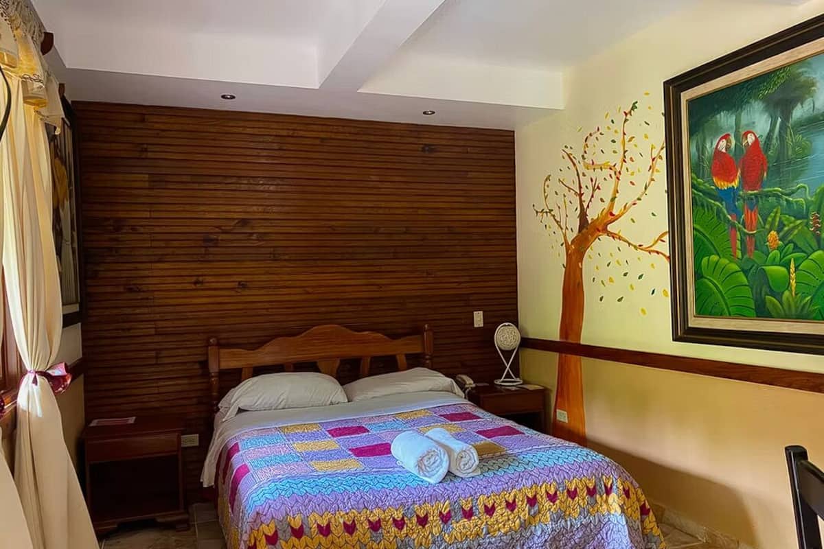 A bedroom with a wooden headboard, a quilted bedspread, a painting of parrots, and a nightstand with a lamp.