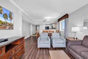 Ein modernes Wohnzimmer mit einem Flachbildfernseher, zwei Sesseln, einem Sofa, einem Holztisch und einem Essbereich mit Küche im Hintergrund.