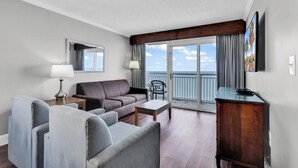 Ett hotellrum med en balkong, en soffa, en fåtölj, ett bord och en tv.