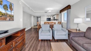 Ett modernt vardagsrum med en platt-TV, två fåtöljer, en soffa, ett träbord och en matplats med ett kök synligt i bakgrunden.