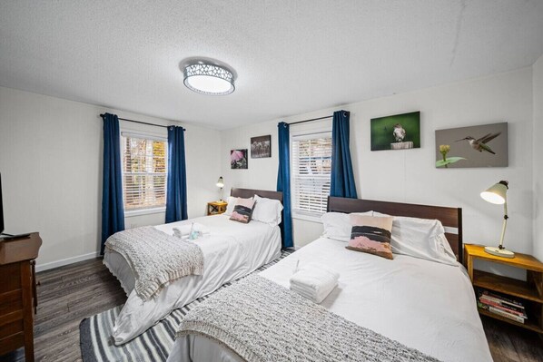 A double room with two beds, a ceiling light, a bedside table with a lamp, a small shelf with books, and a window with curtains.