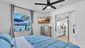 A bedroom with a bed, a ceiling fan, a window with curtains, a view of the outside, and a kitchen area with cabinets and a refrigerator. - Spacious Family Getaway Near AMI Beaches! Bikes, Beach Gear, Pets & Sleeps 8 (Bradenton)