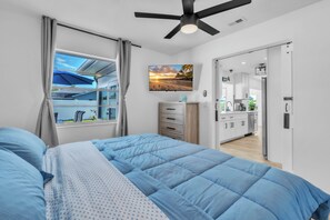 Ein Schlafzimmer mit Bett, Deckenventilator, Fenster mit Vorhängen, Blick ins Freie und einer Küchenzeile mit Schränken und Kühlschrank.