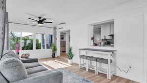 A modern living room with a grey sofa, a small bar area with stools, and a view of the outdoors through large windows. - Spacious Family Getaway Near AMI Beaches! Bikes, Beach Gear, Pets & Sleeps 8 (Bradenton)