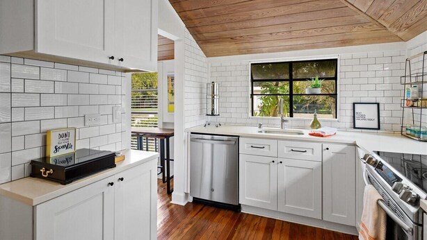 A modern kitchen with white cabinetry, a wooden ceiling, and a window with a view of greenery.