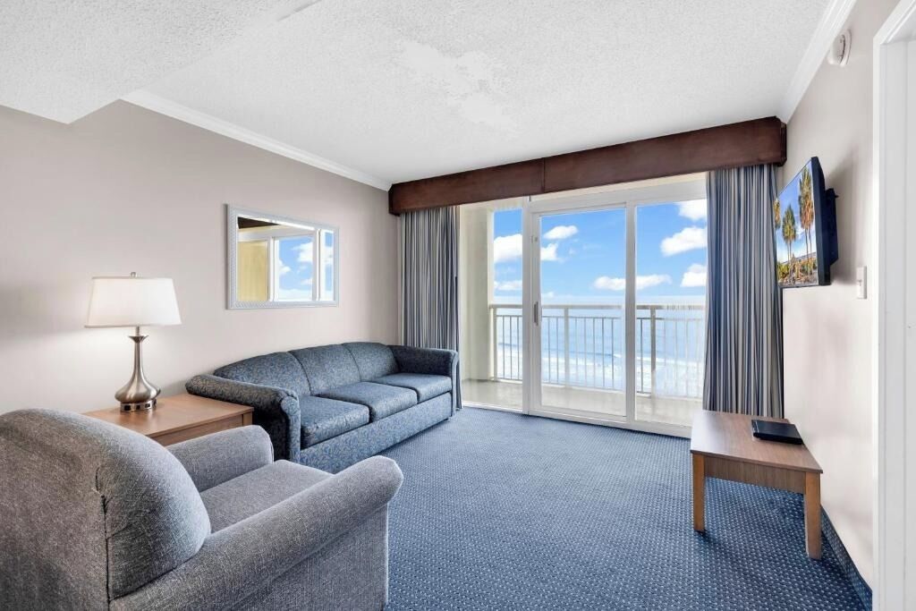 A hotel room with a sofa, a small table, a lamp, a TV, and a balcony with a view.