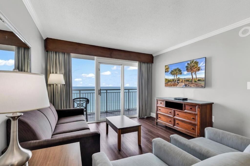 A hotel room with a balcony, a sofa, a table, a TV, and a wooden dresser.