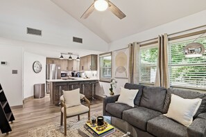 Un salon moderne avec un canapé gris, une table basse sur laquelle sont posés des livres et une tasse, un plancher en bois, une cuisine en arrière-plan et un ventilateur de plafond.