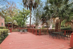 Terraza de madera roja con mobiliario exterior, rodeada de árboles y edificios.