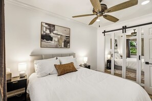 Une chambre à coucher comprenant un lit, des tables de chevet, un ventilateur de plafond, un tableau abstrait accroché au mur et un placard à portes coulissantes.