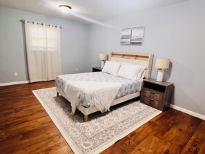 Un dormitorio con cama, mesitas de noche, una alfombra, una ventana con cortinas y una obra de arte colgada en la pared.