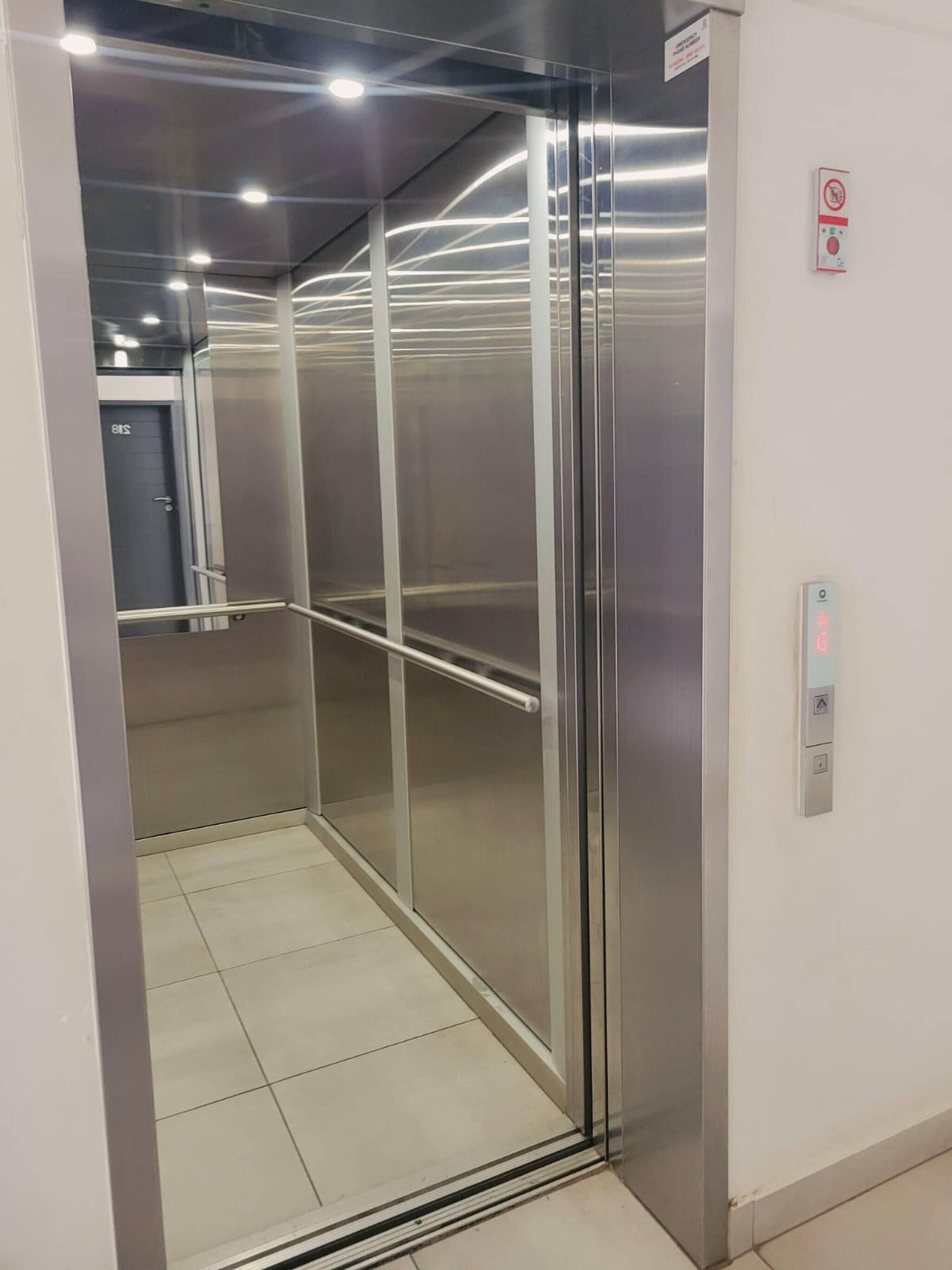 Un ascensor modern amb parets metàl·liques i una porta de vidre, equipat amb un panell de control i un botó d'aturada d'emergència.