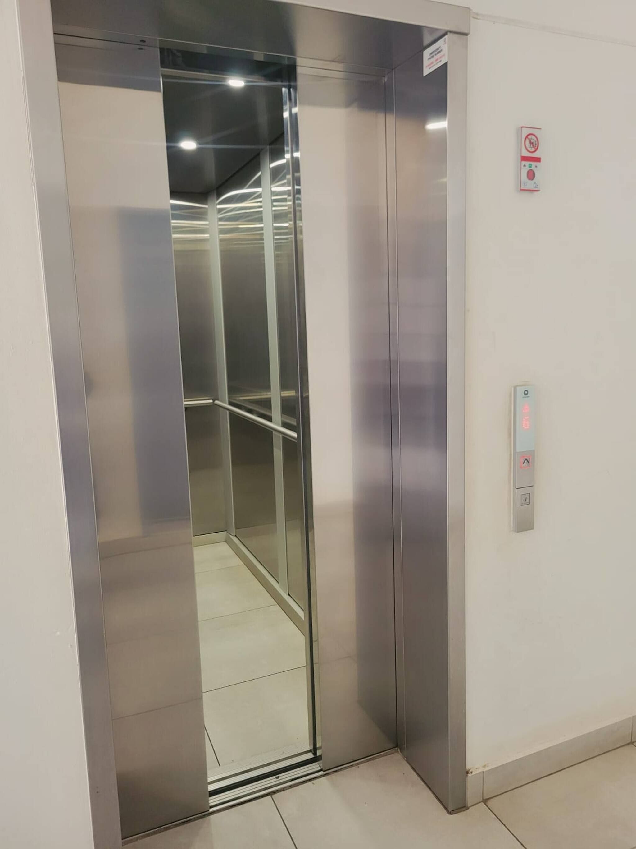 An elevator with metallic walls and a glass door, a control panel with buttons, and a no-smoking sign on the wall.