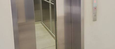 An elevator with metallic walls and a glass door, a control panel with buttons, and a no-smoking sign on the wall.
