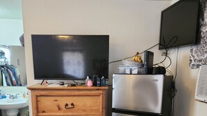A kitchen with a wooden chest of drawers, a stainless steel refrigerator, a flat-screen TV, and a microwave.