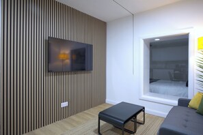 Ein modernes Wohnzimmer mit einem flachbildfernseher an einer Holzwand, einem kleinen schwarzen Tisch, einem grauen Sofa mit gelbem Kissen und einem großen Fenster mit Blick auf ein Bett.