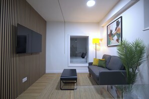 Ett modernt vardagsrum med en platt-TV, en soffa med kuddar, ett soffbord i glas, ett stort fönster och en golvlampa.
