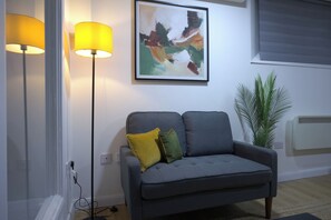 En grå soffa med två kuddar, en golvlampa, en tavla, en krukväxt och ett element.