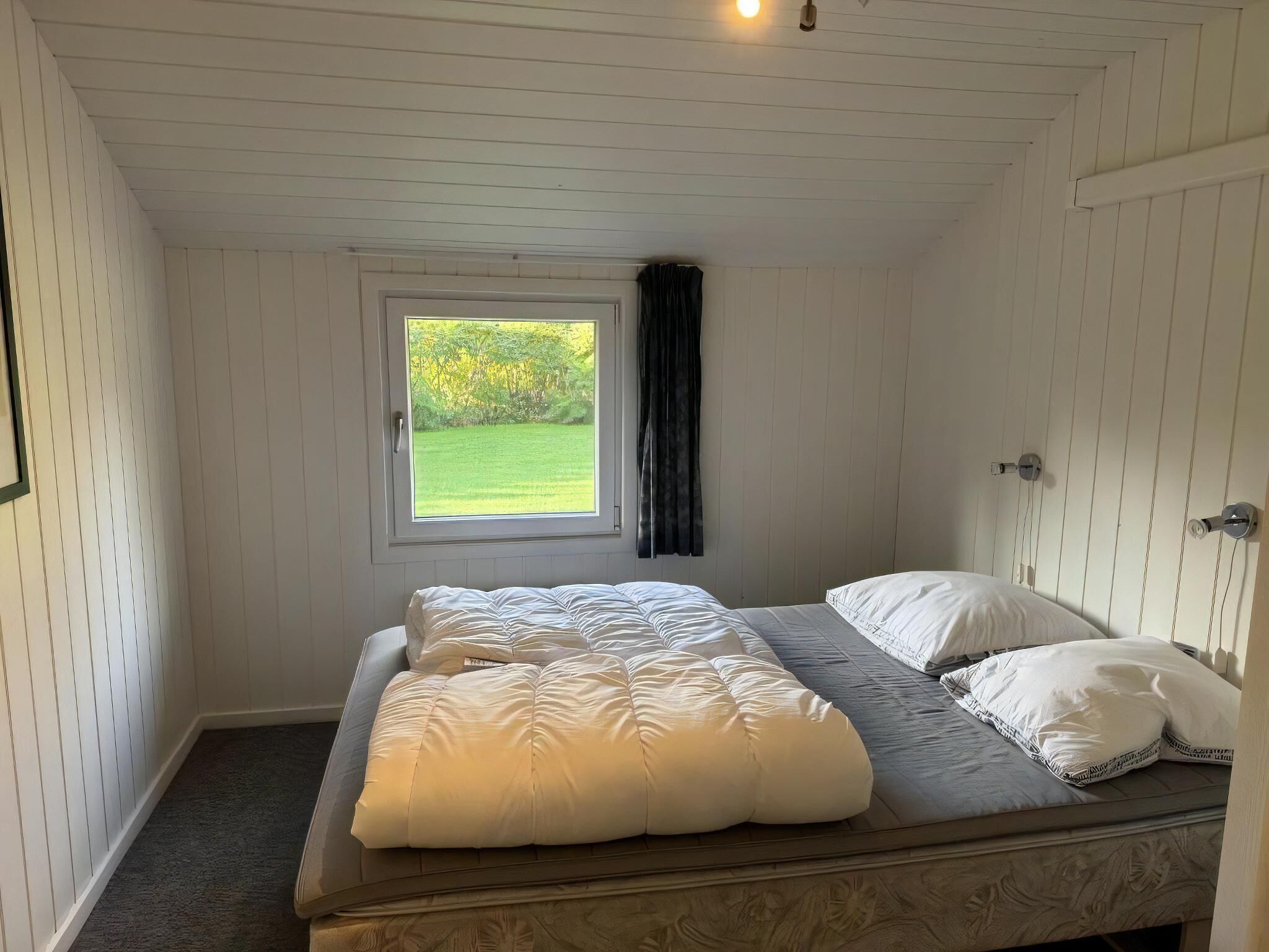 Uma cama de solteiro com cabeceira, uma janela com vista para o verde e teto branco.