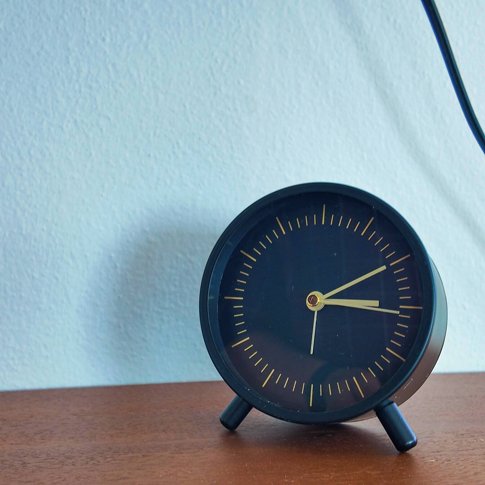 Une horloge analogique noire, avec des aiguilles des heures et des minutes dorées, posée sur une surface en bois devant un mur uni.