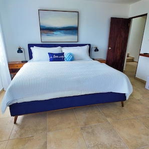 A bedroom with a large bed, two bedside tables, a nightstand, a door, and a painting on the wall. - Casa Las Nubes-cayey (Cayey)