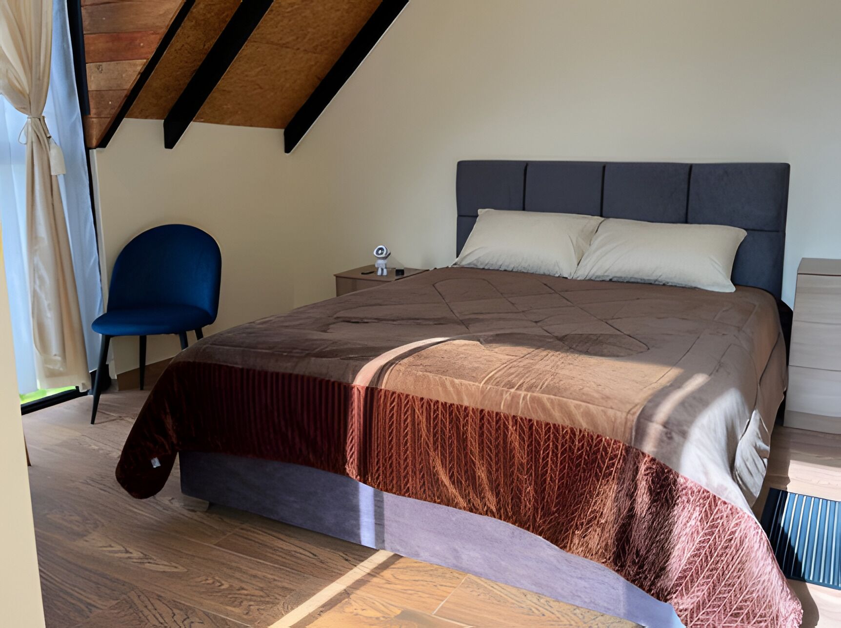 Un dormitorio con cama, una silla, una mesita de noche y una ventana con cortinas.