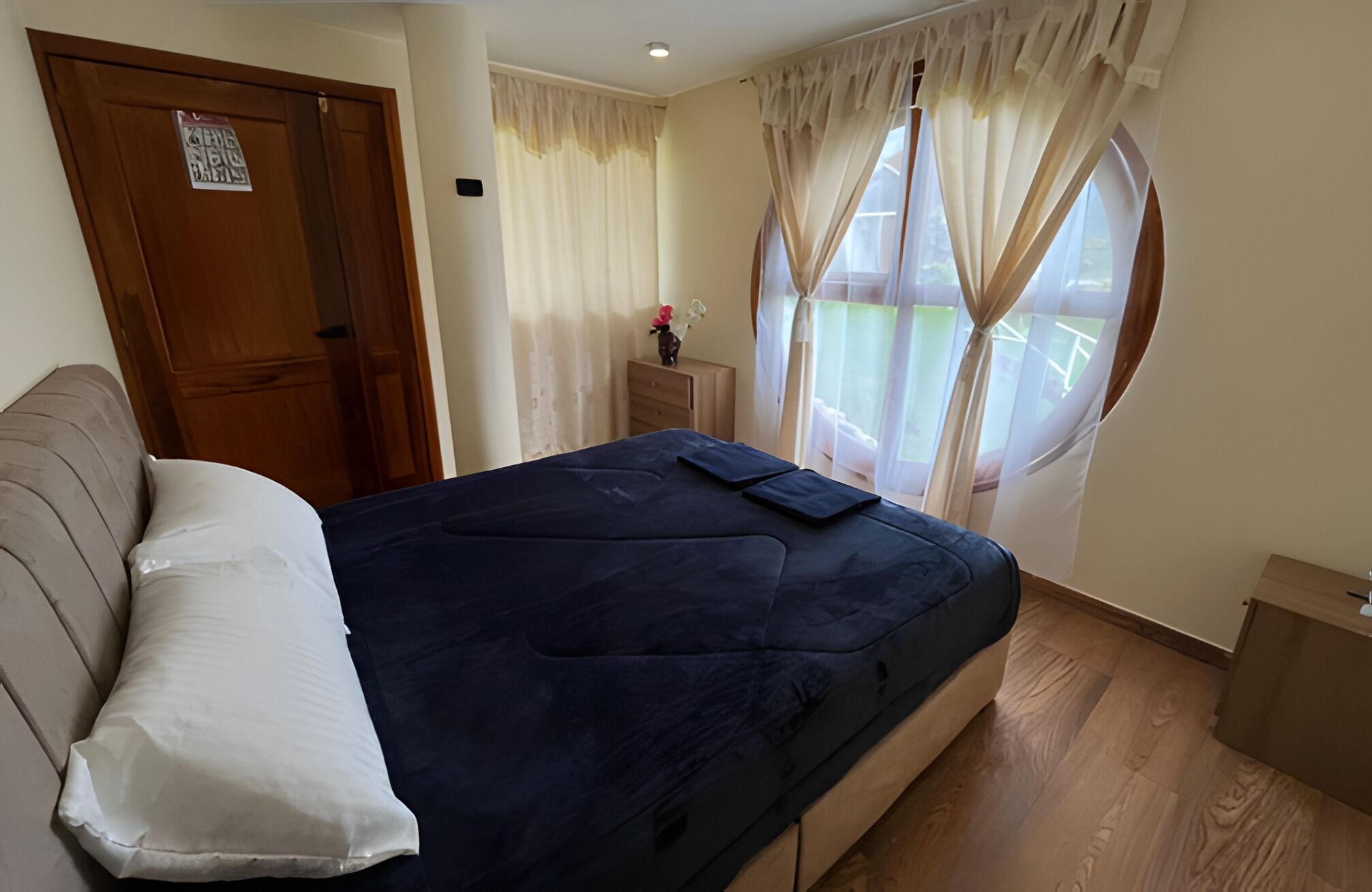 Un dormitorio con una cama, una puerta de madera, una mesita con un florero y un ventanal con cortinas.