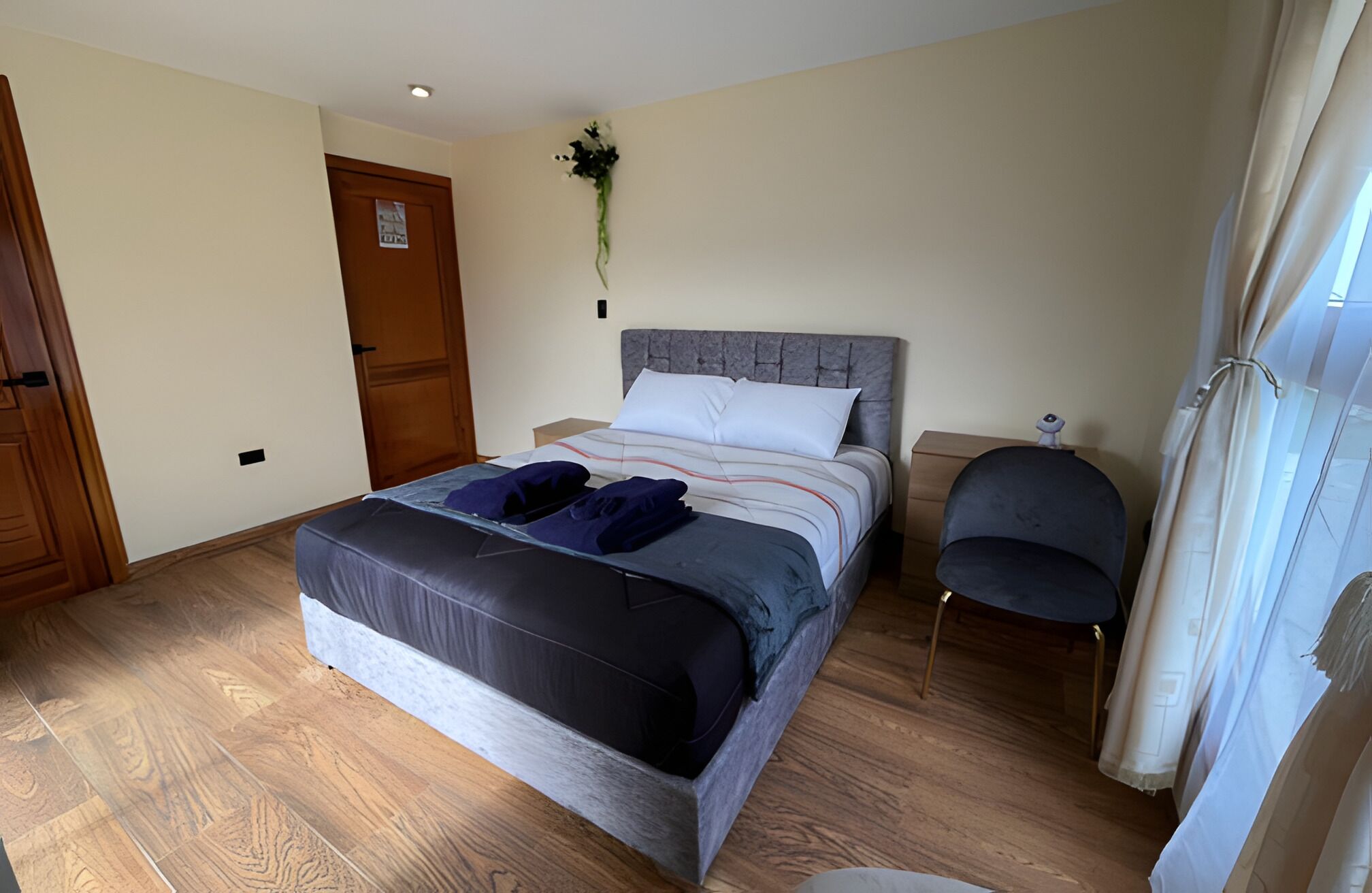 A bedroom with a bed, a chair, a nightstand, a door, and a wall-mounted light fixture.