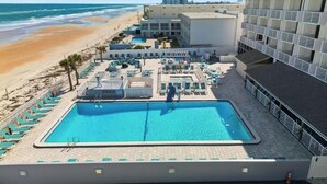 Una piscina en la azotea con vistas a la playa y al océano, un área de descanso con tumbonas, y un edificio con fachada de diseño geométrico.