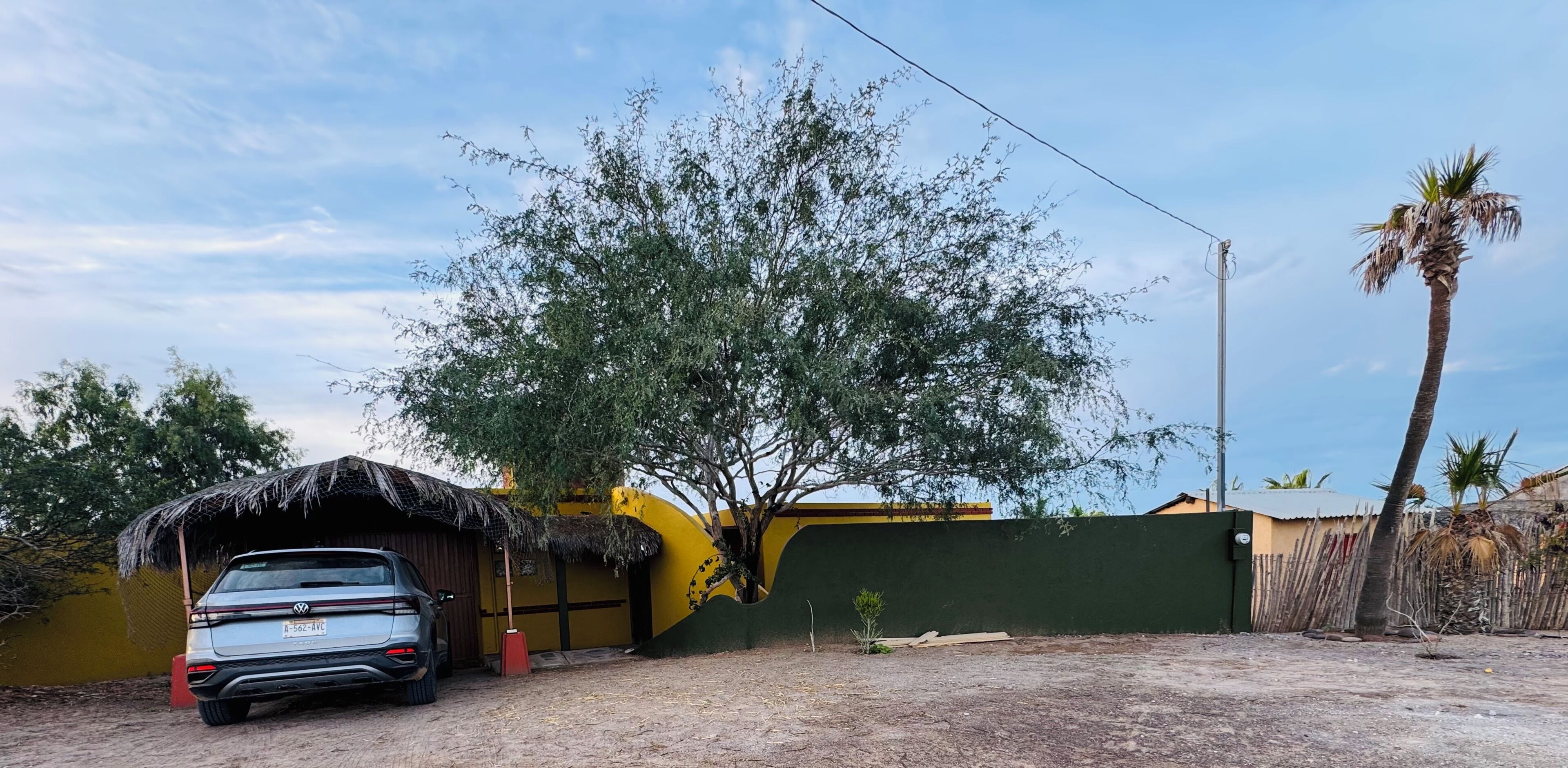 A car parked in front of a building with a thatched roof, a palm tree, and a fence.