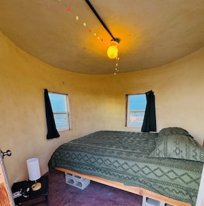 A bedroom with a curved ceiling, two windows with curtains, a bed with patterned bedding, a nightstand with a lamp, and a view of the outside.