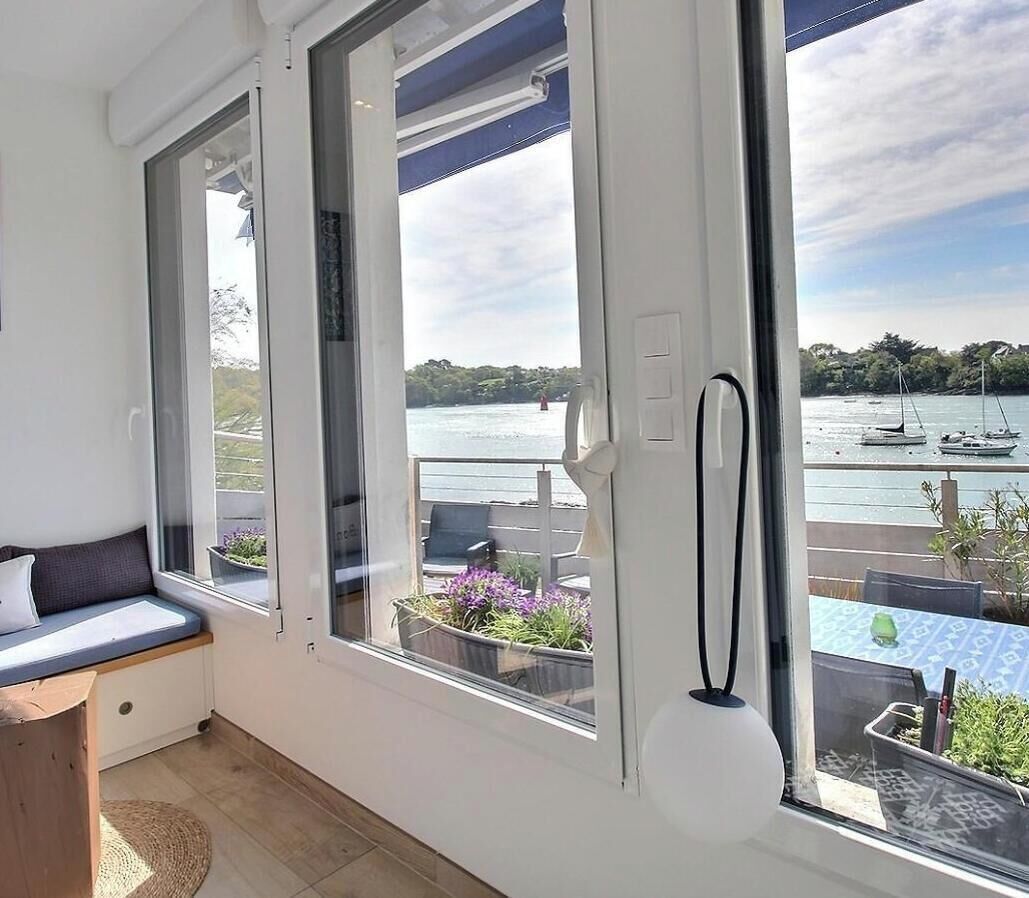 A room with a large window overlooking a body of water, a balcony with outdoor seating, and a view of boats.